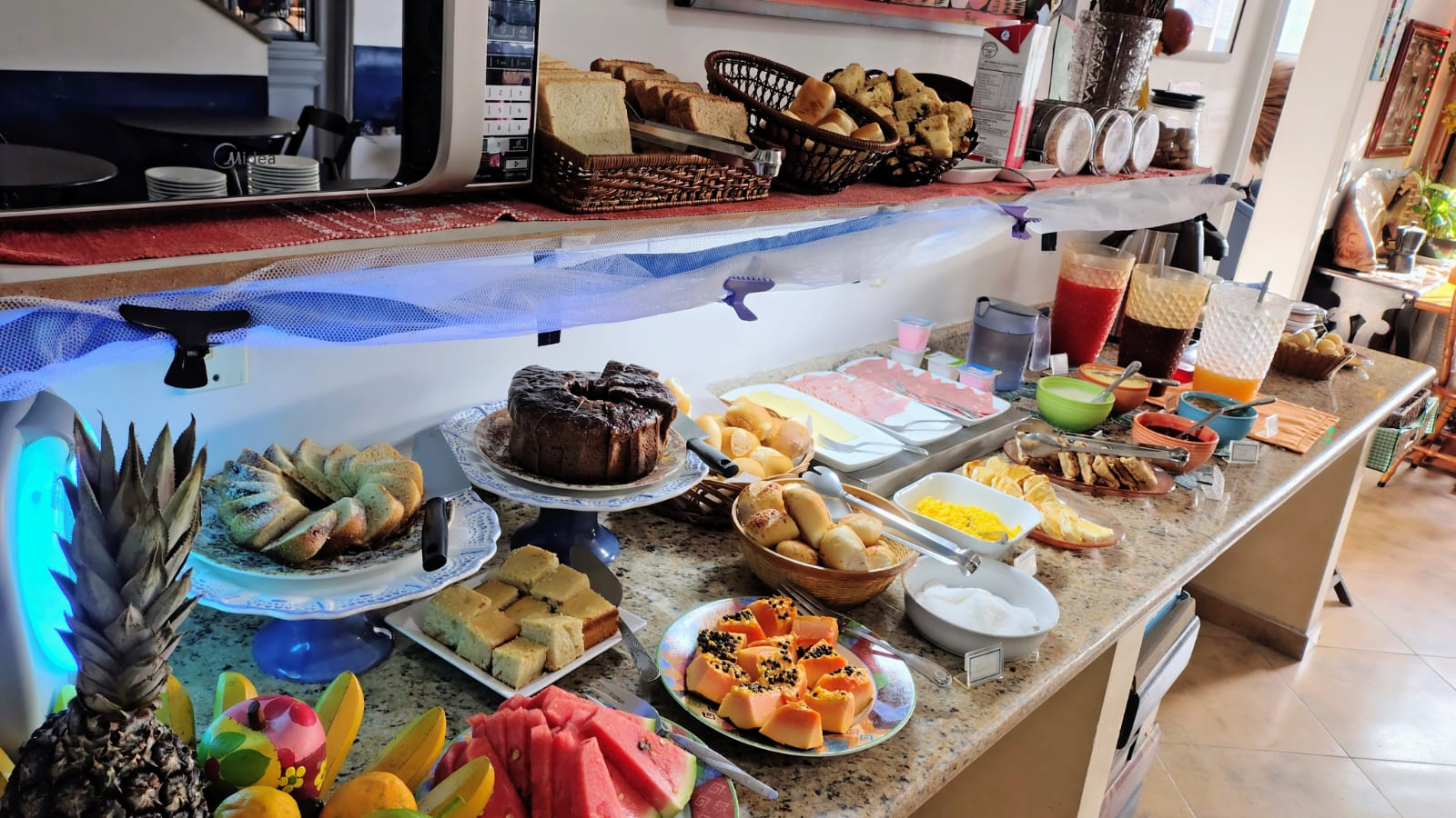 Buffet de caf&eacute; da manh&atilde; montado sobre balc&atilde;o de granito, com bolos (inclusive bolo de chocolate), fatias de p&atilde;o e p&atilde;es variados em cestos, frutas frescas como melancia, mam&atilde;o e abacaxi, sucos em jarras, frios (queijo e presunto), ovos mexidos, sandu&iacute;ches, manteiga e acompanhamentos, al&eacute;m de micro-ondas e utens&iacute;lios ao fundo em ambiente iluminado e organizado.