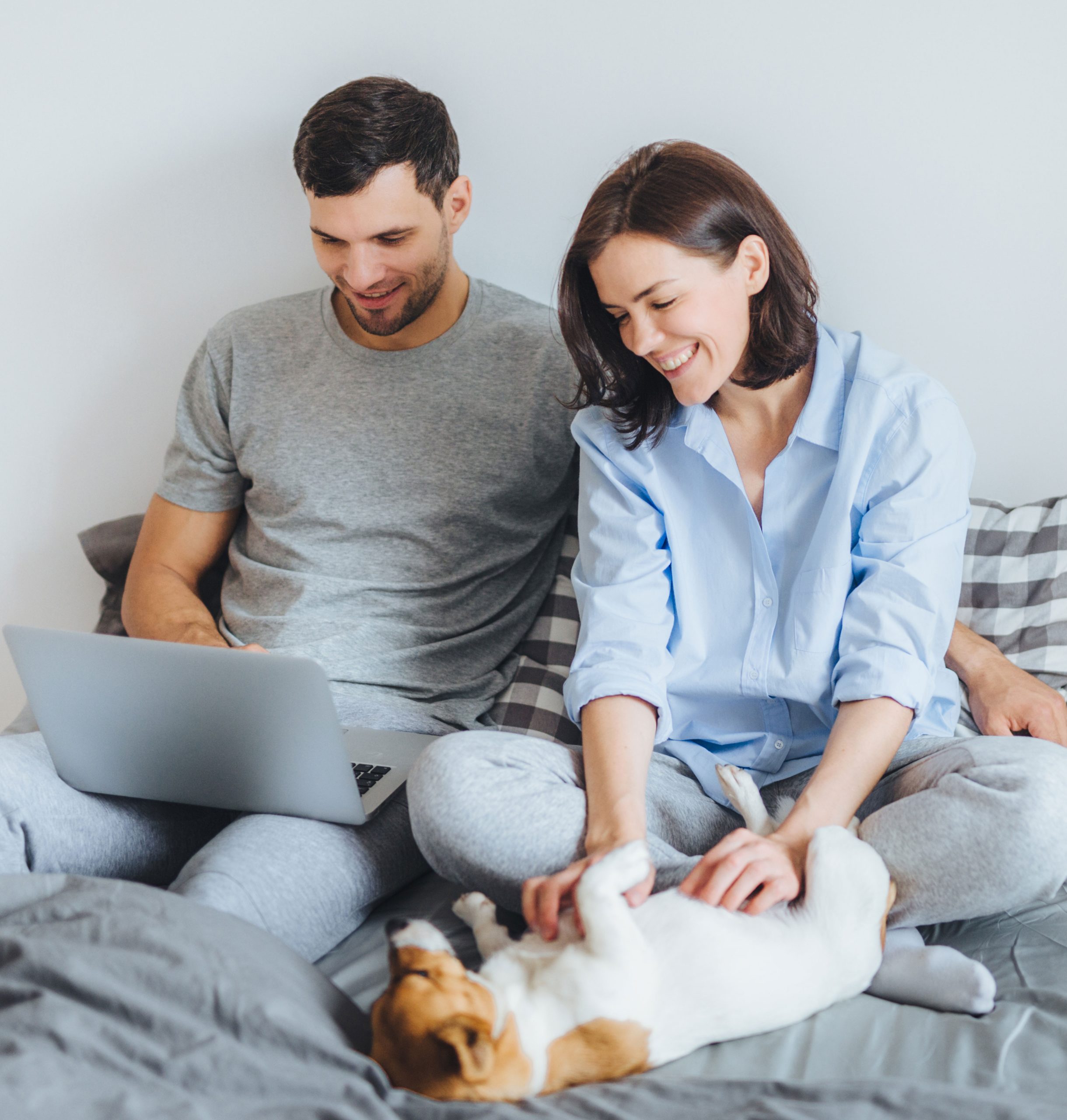Casal sentado na cama em um ambiente claro e aconchegante, sorrindo enquanto usa um notebook e faz carinho em um cachorro deitado de barriga para cima entre eles.