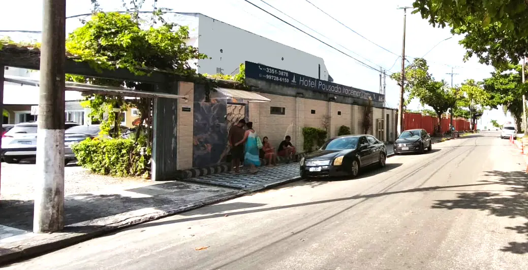 Fachada do hotel vista da rua em um dia ensolarado, com muro claro e port&atilde;o de entrada aberto, placa com o nome do hotel na parte superior, algumas pessoas sentadas pr&oacute;ximas &agrave; entrada e carros estacionados ao longo da via arborizada.