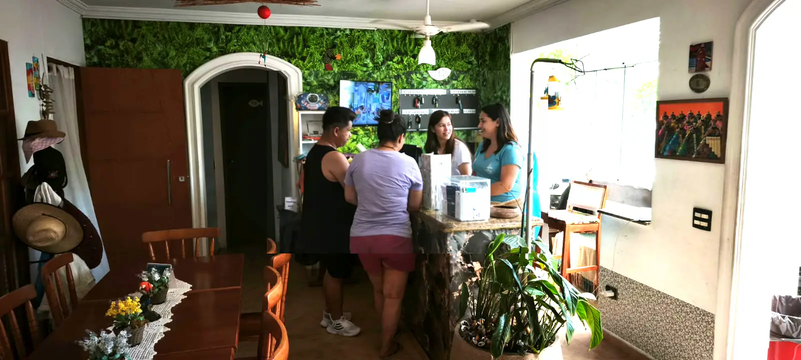 Interior de recep&ccedil;&atilde;o aconchegante com parede verde decorativa ao fundo, onde duas mulheres atendem atr&aacute;s de um balc&atilde;o de pedra enquanto um casal de  h&oacute;spedes &eacute; atendido; &agrave; esquerda h&aacute; mesa de madeira com toalha rendada e flores, e &agrave; direita uma janela ampla iluminando o ambiente.