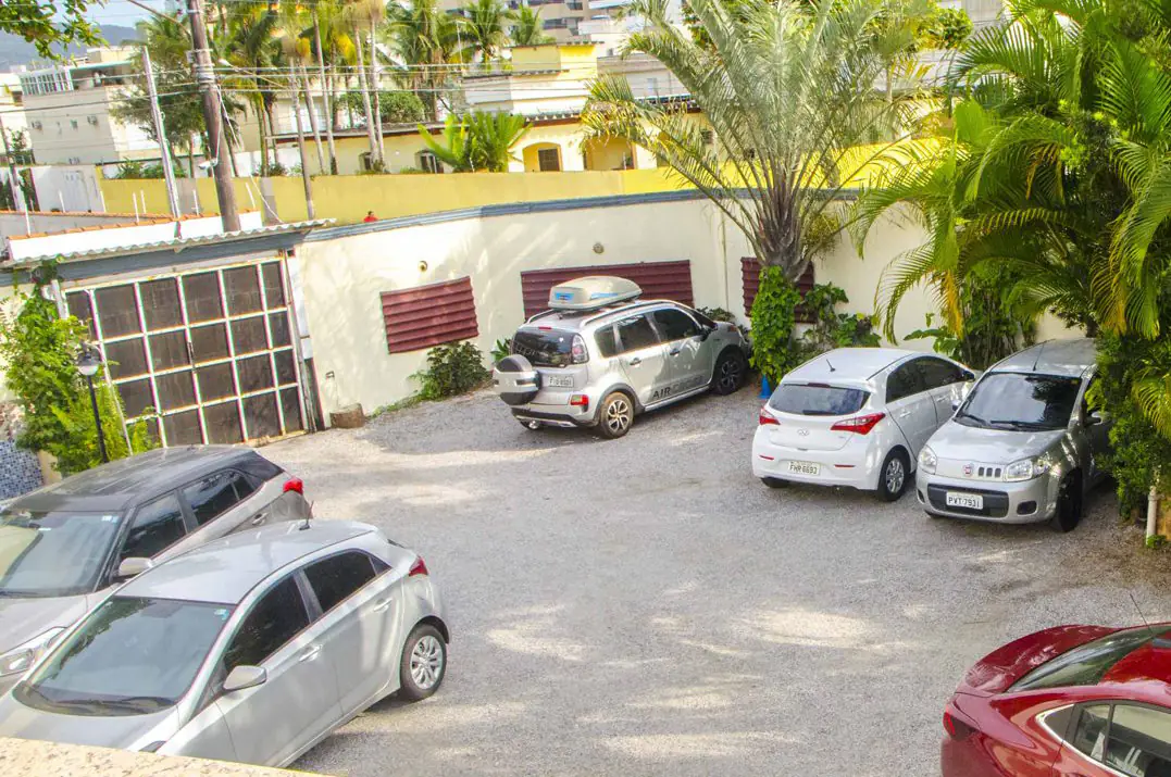 Estacionamento externo com piso de brita, cercado por muros e vegeta&ccedil;&atilde;o tropical, com v&aacute;rias vagas ocupadas por carros, palmeiras ao redor e casas residenciais ao fundo em &aacute;rea ensolarada.